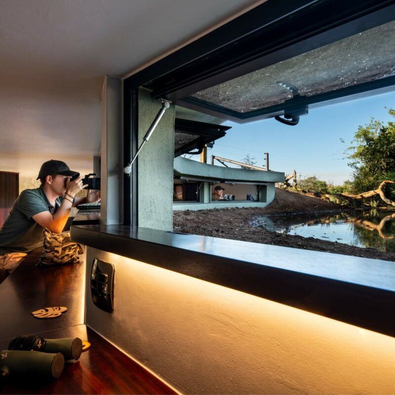 Man photographing wildlife from inside a lodge, overlooking a water source.