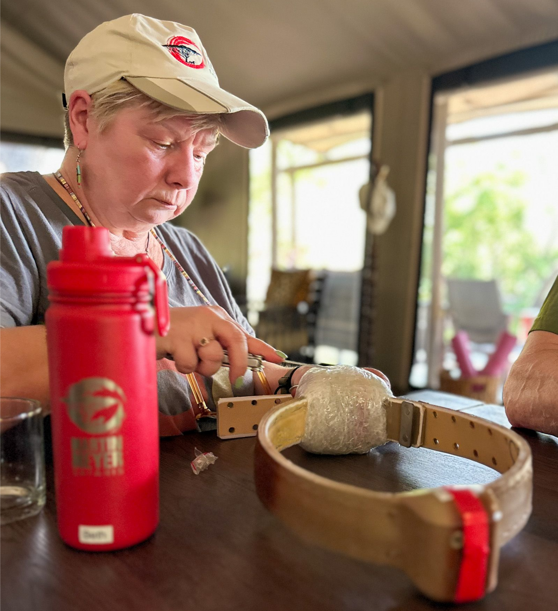 Martin Meyer Safari guest with a cap working on a Elephant collar at a table, red water bottle beside her.