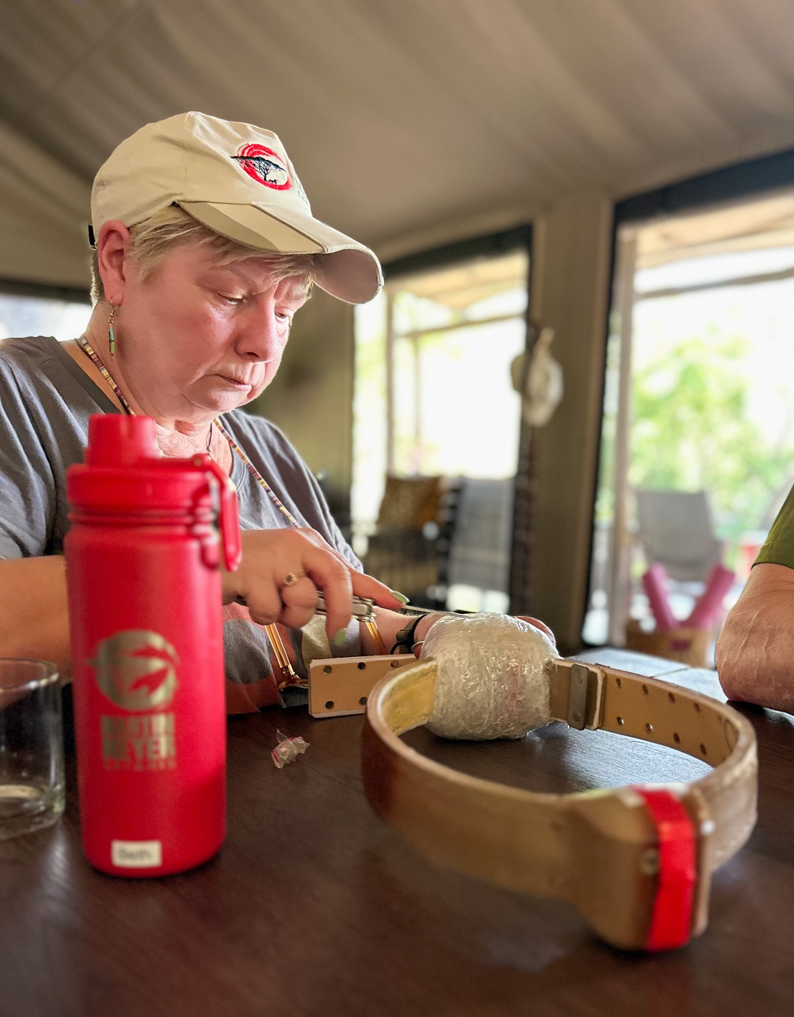 Woman cutting something on a table, with a red water bottle and a dog collar. Inside, natural light.