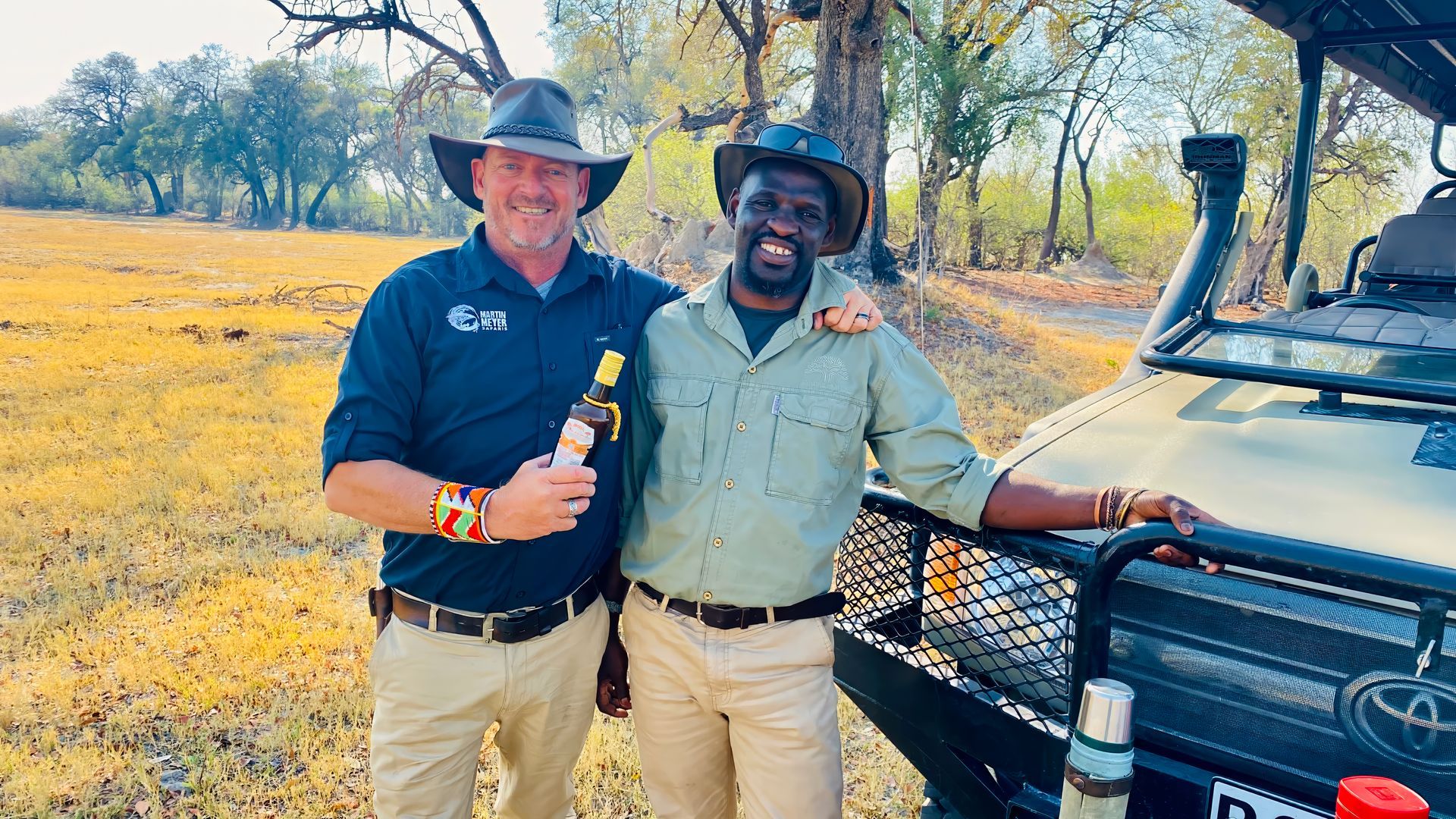 Two men smiling, posing outdoors with a safari vehicle; one holds a drink.