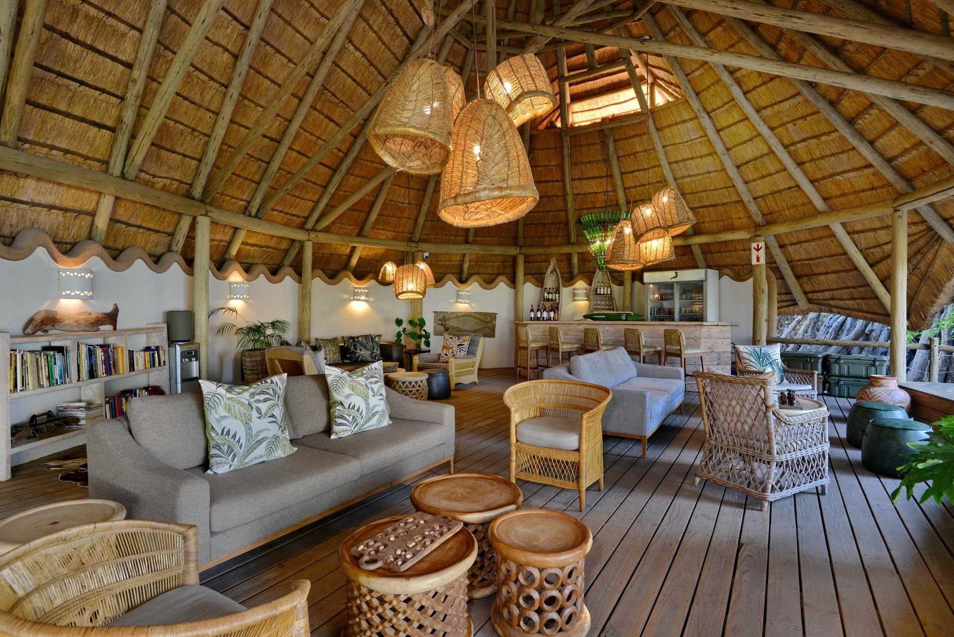 A relaxing lounge area with woven furniture, thatched roof, and hanging lamps. Features a bar and bookshelves.