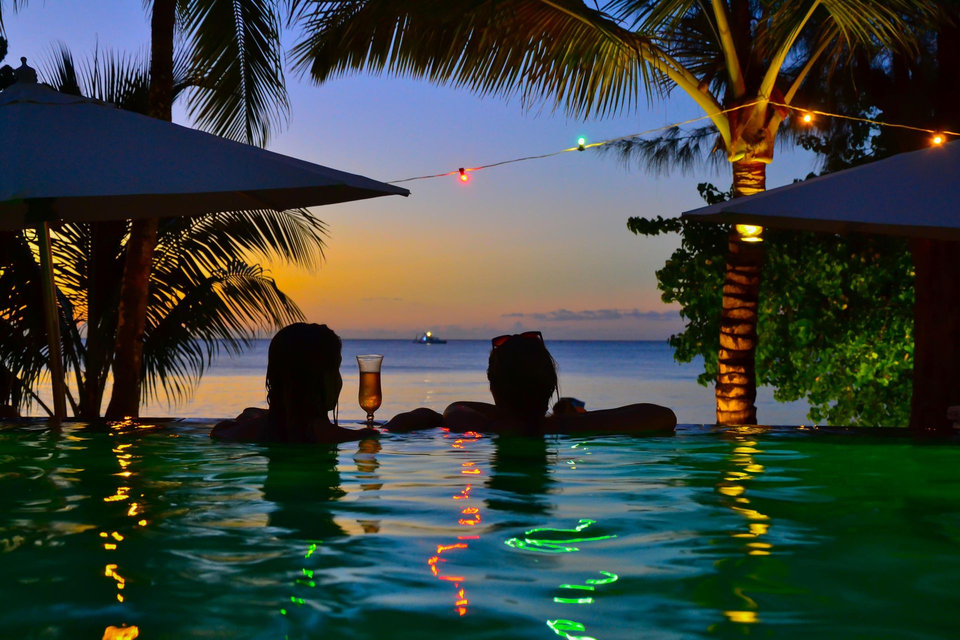 Two people in pool at sunset, looking out over ocean, with drinks. Palm trees, umbrellas and lights.