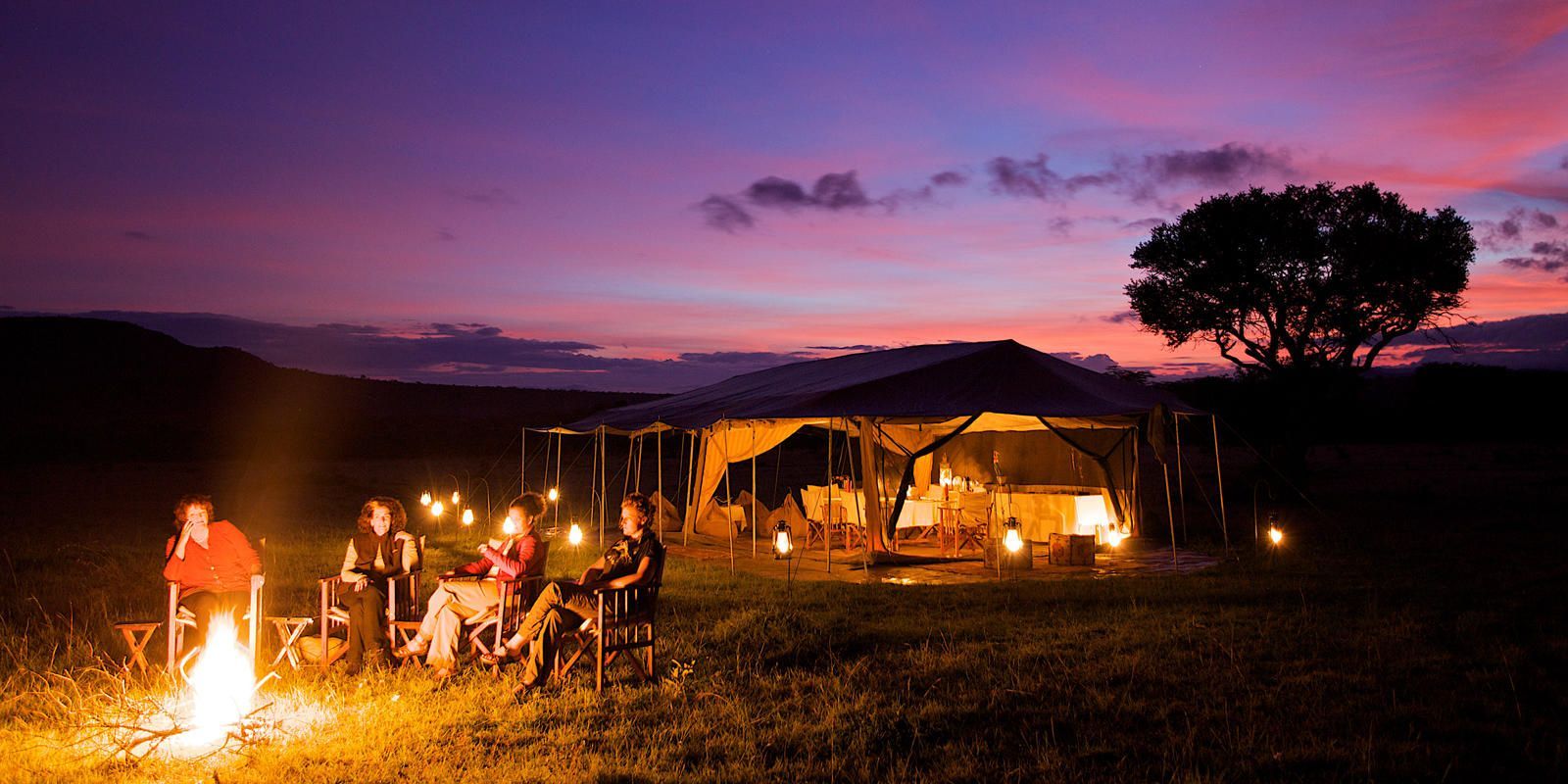 Campers gather around a campfire at dusk, with a tent in the background, under a colorful sky.