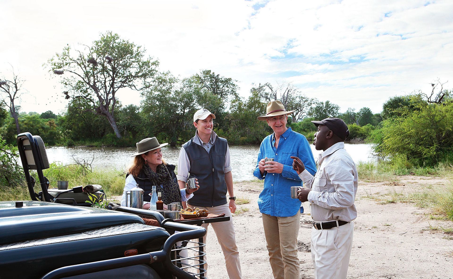 Four people in safari attire converse near a vehicle and a river in a sunny setting.