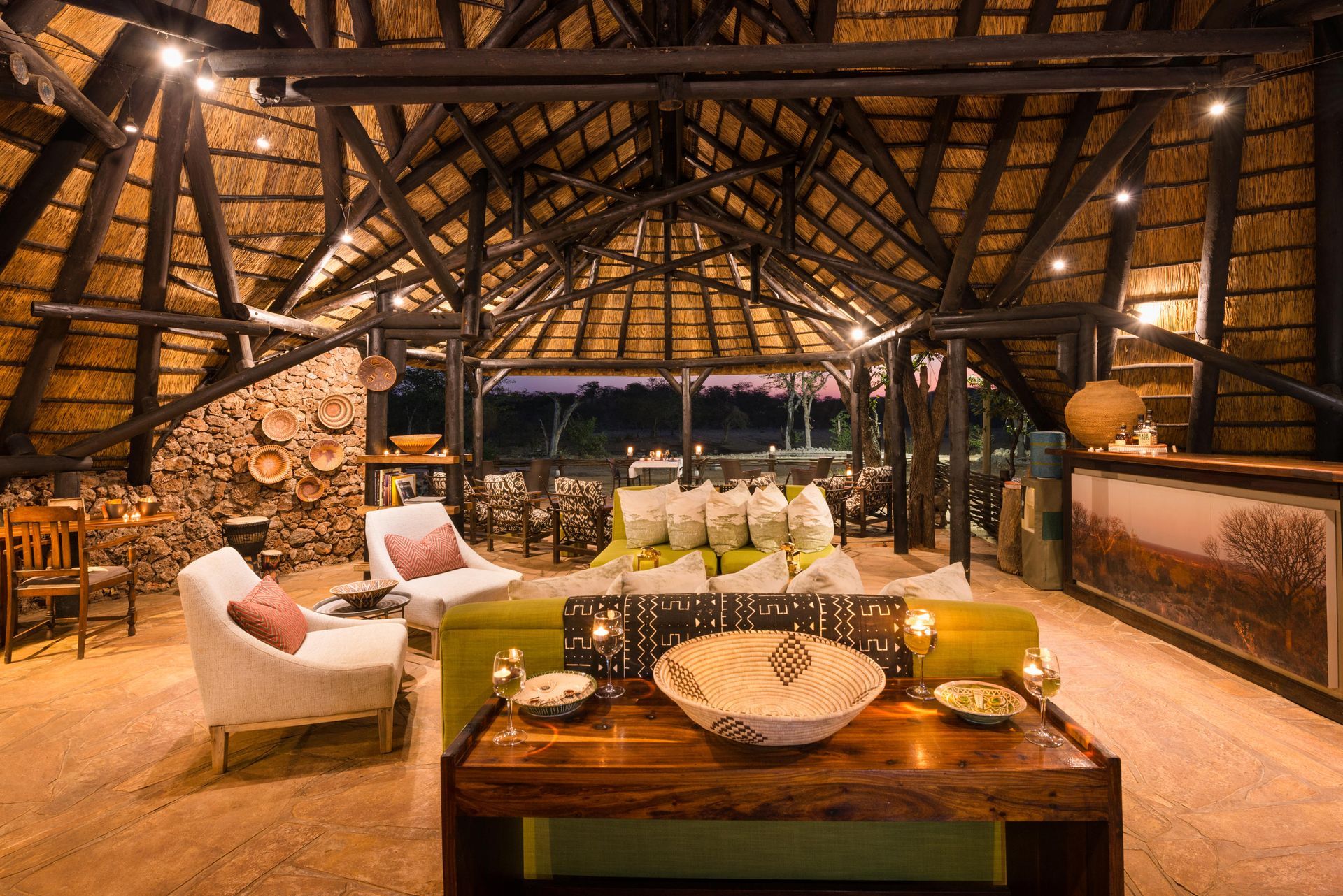 Cozy interior with thatched roof and wooden beams. Furniture includes white sofas, a wooden table, and a bar area.