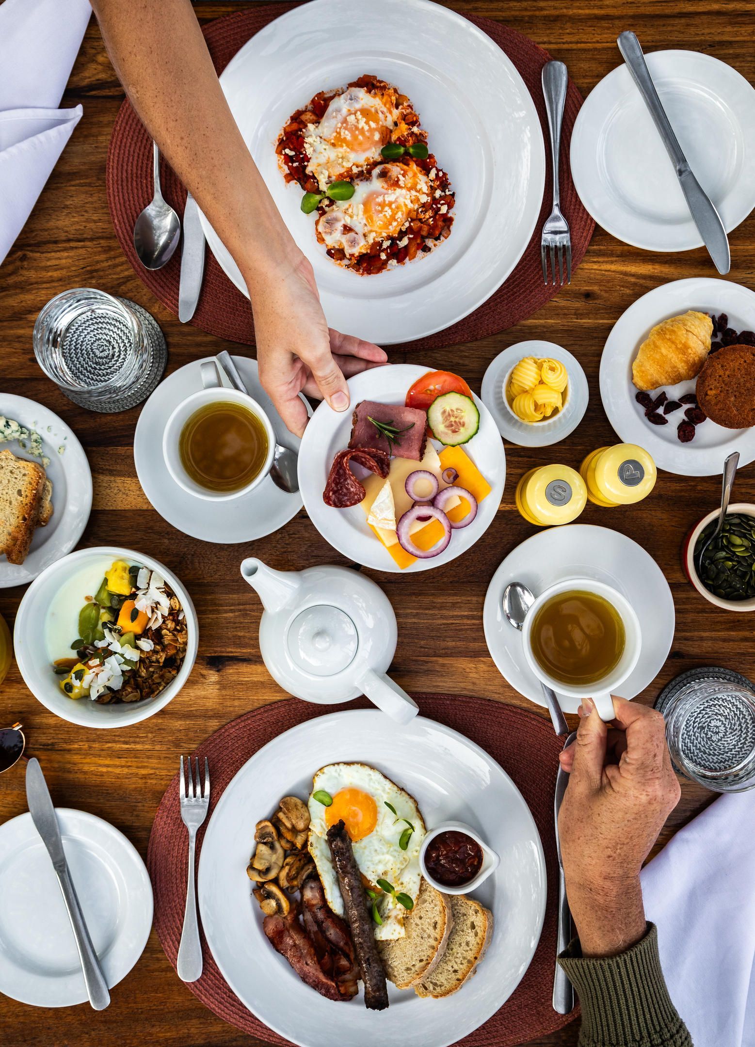 Overhead shot of breakfast on a wooden table, hands reaching for plates of food, including eggs and fruit.