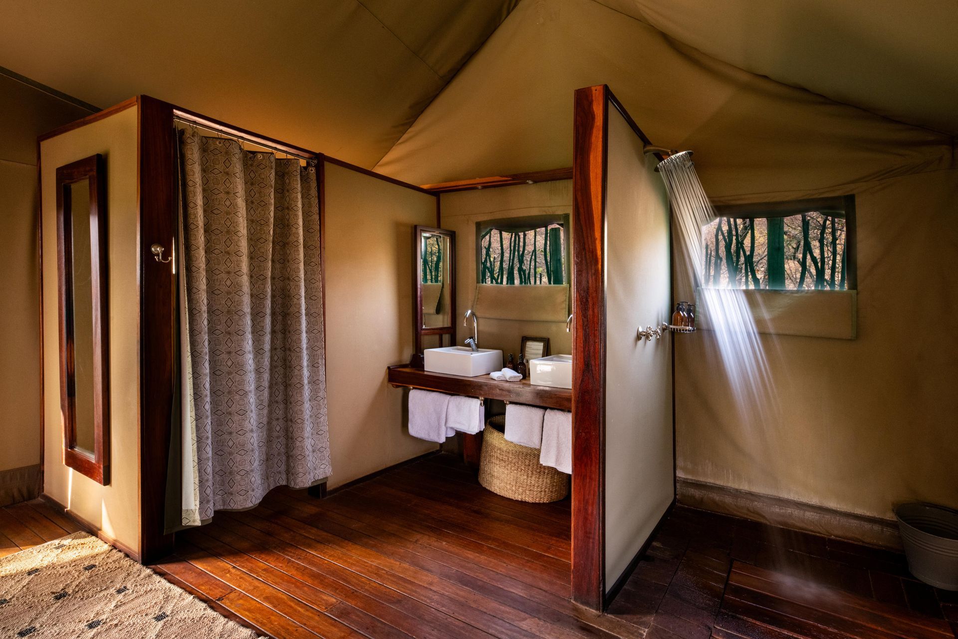 Safari camp bathroom. Wooden floor, open shower, sinks, and a curtain-covered space. Sunlight streams in through windows.