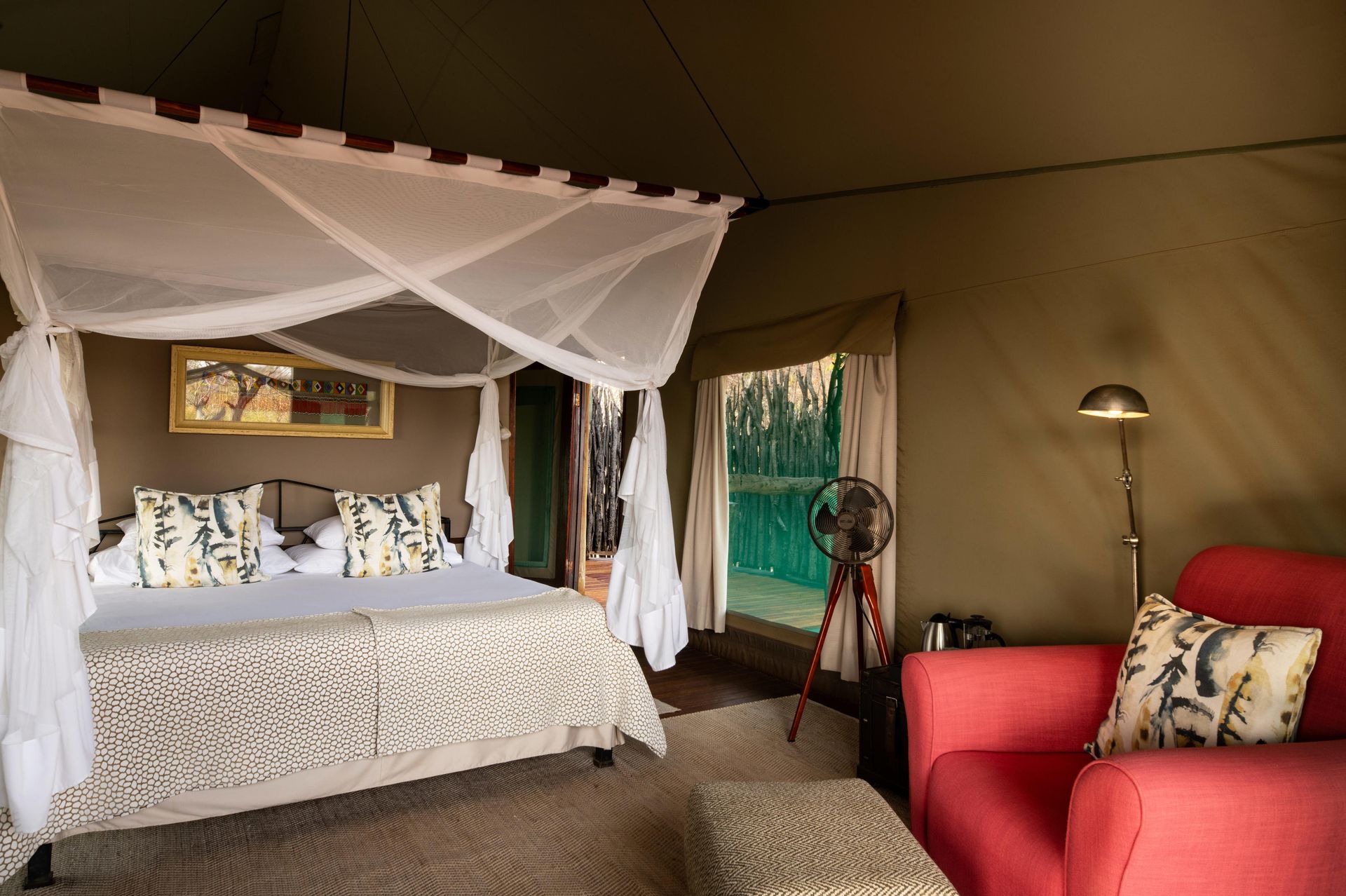 Interior of a safari tent with a bed, red armchair, and open door looking onto a view.