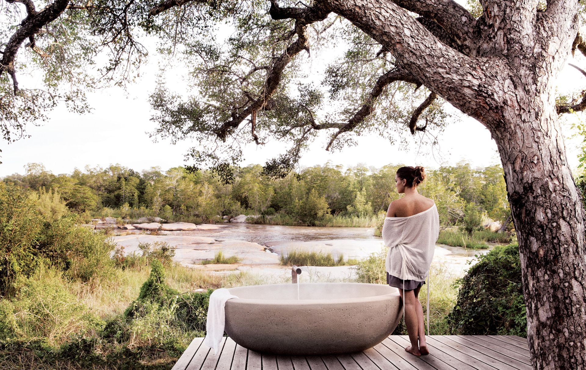 Woman stands near an outdoor bathtub, overlooking a river and trees.