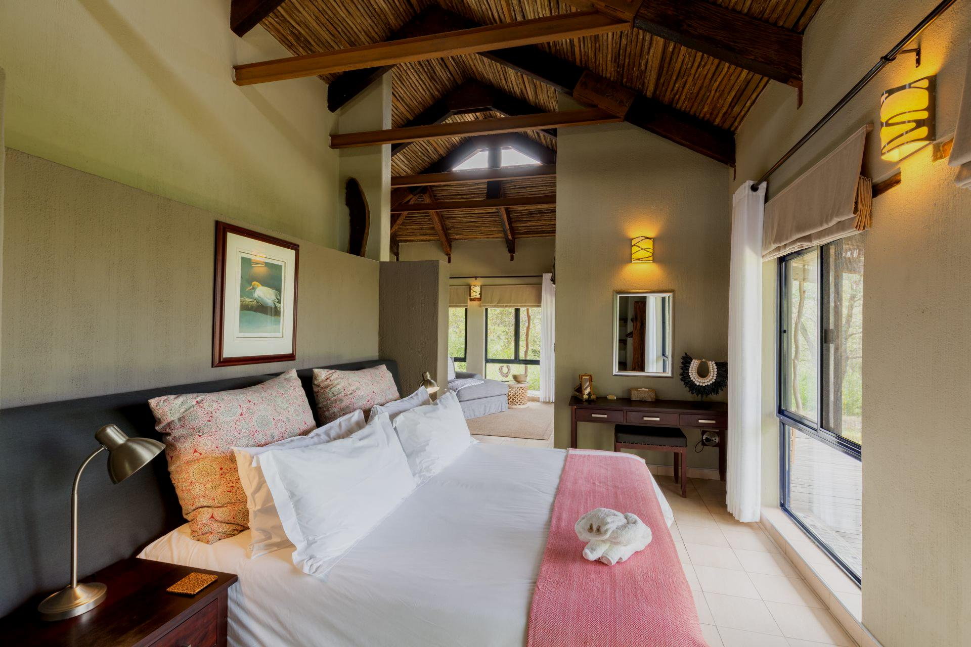 Bedroom with a king-size bed, featuring a pink blanket, under a thatched roof. Adjacent to a window with view.