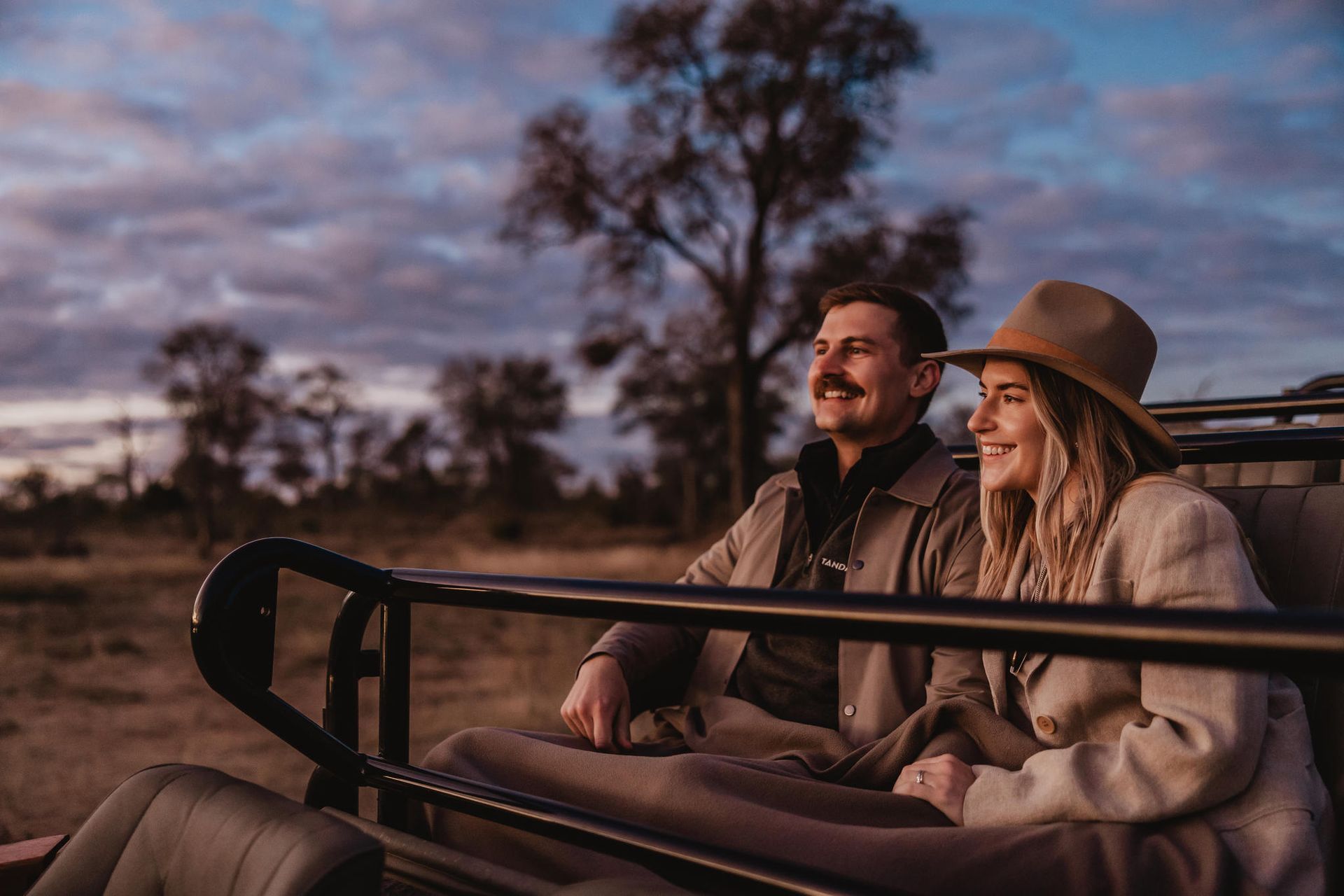 Couple on safari, smiling in jeep at sunset.