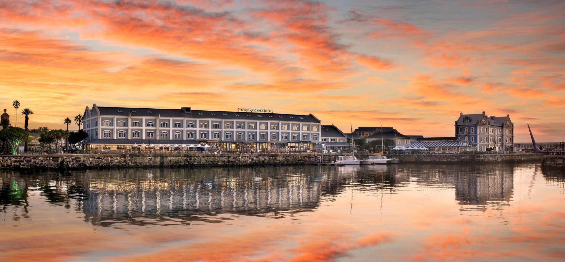 Waterfront hotel at sunset, reflecting in calm water. Orange sky.