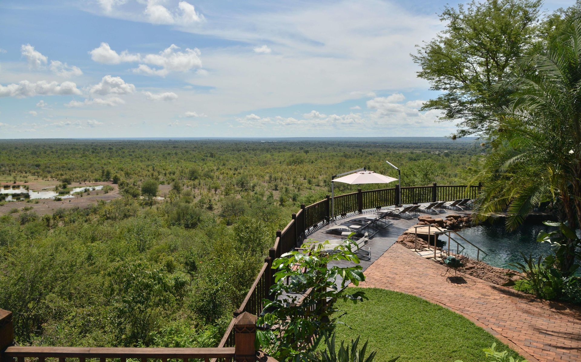 Overlook of a lush green landscape with a pool and seating area, under a bright blue sky dotted with white clouds.