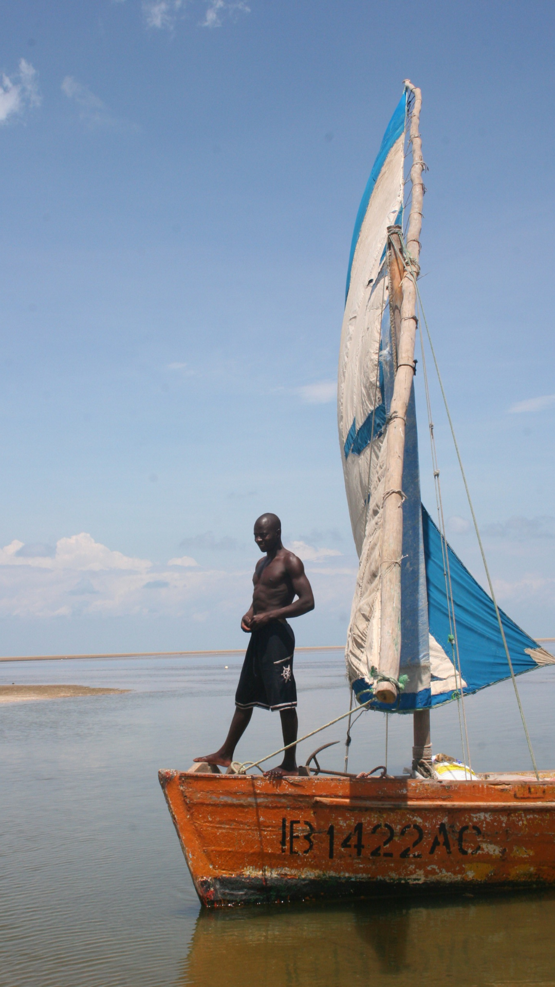 Man on a sailboat, brown boat, blue and white sail, on calm water, sunny day.