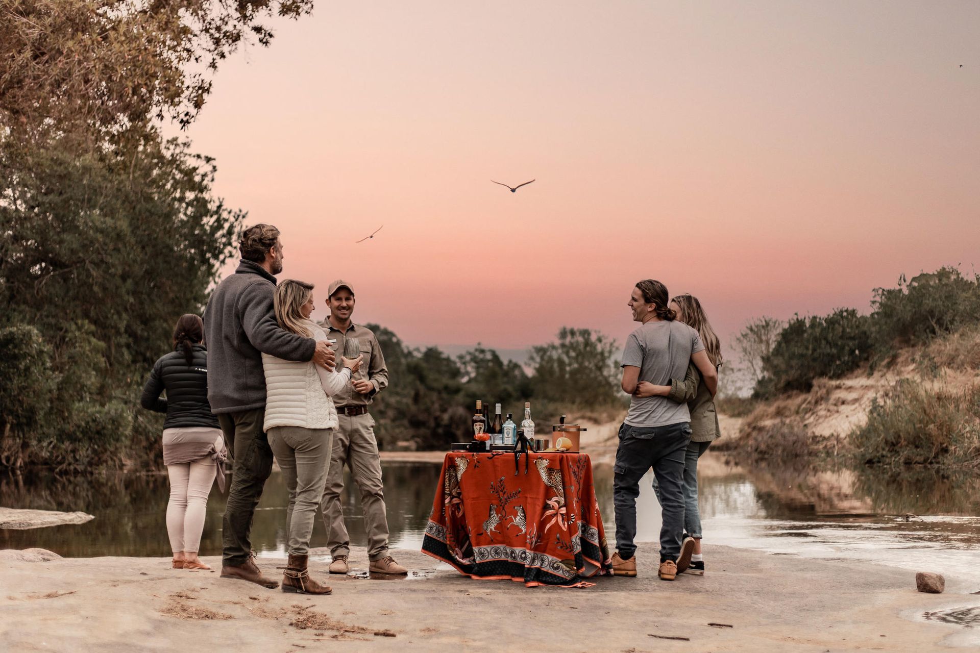 People by a riverside table at sunset, enjoying drinks, embraced.
