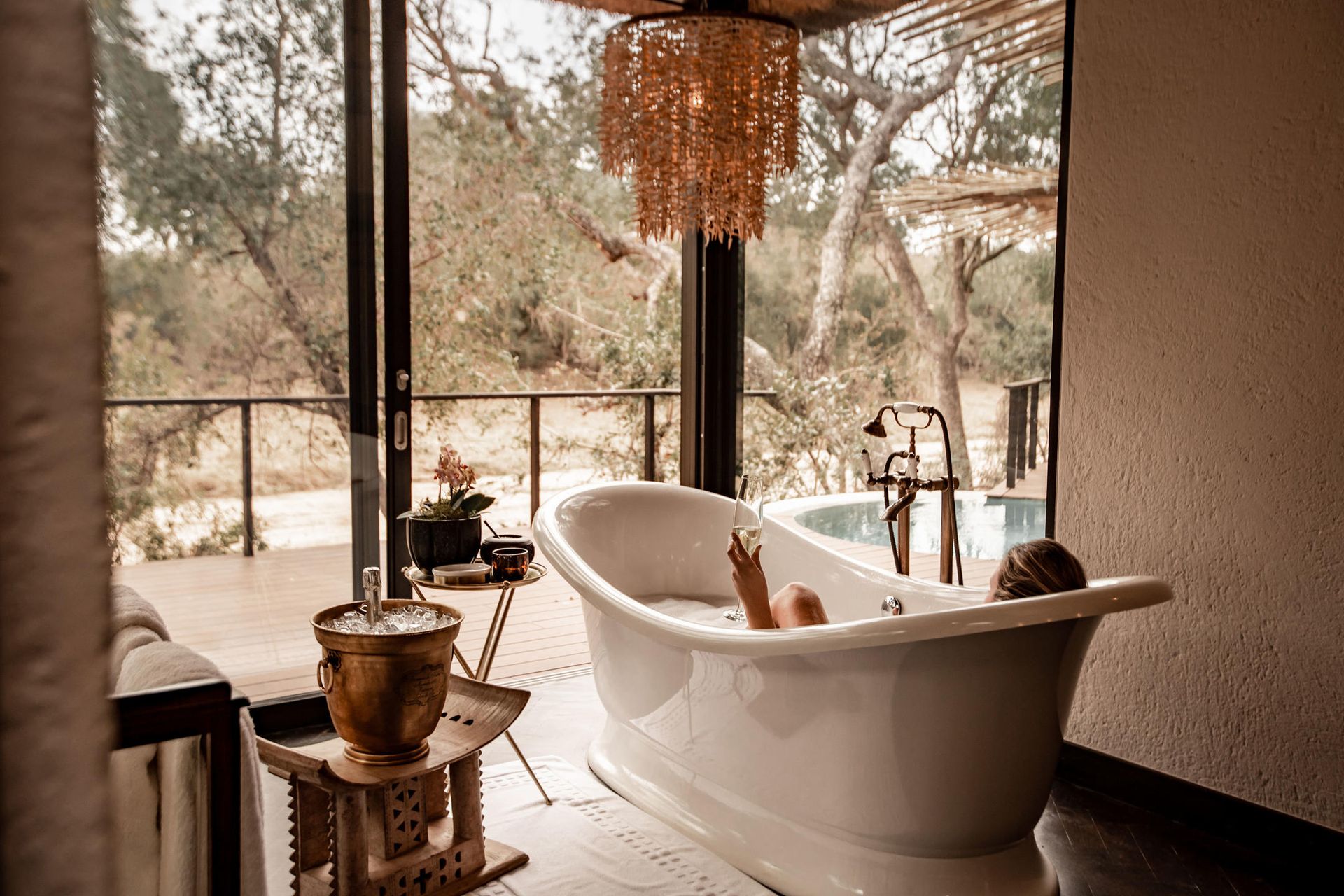 Woman in a clawfoot tub, arm raised, with view of trees, drink beside her.