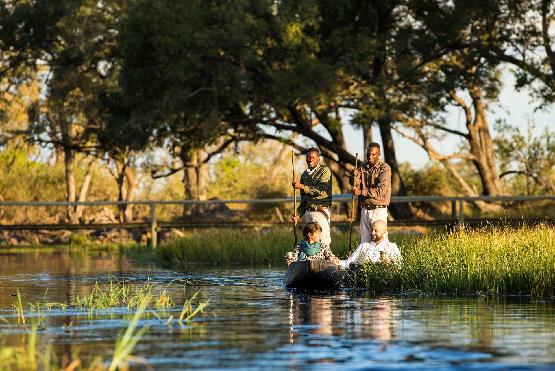 People in a small boat navigate a calm river, guided by two men with poles, near greenery and trees.