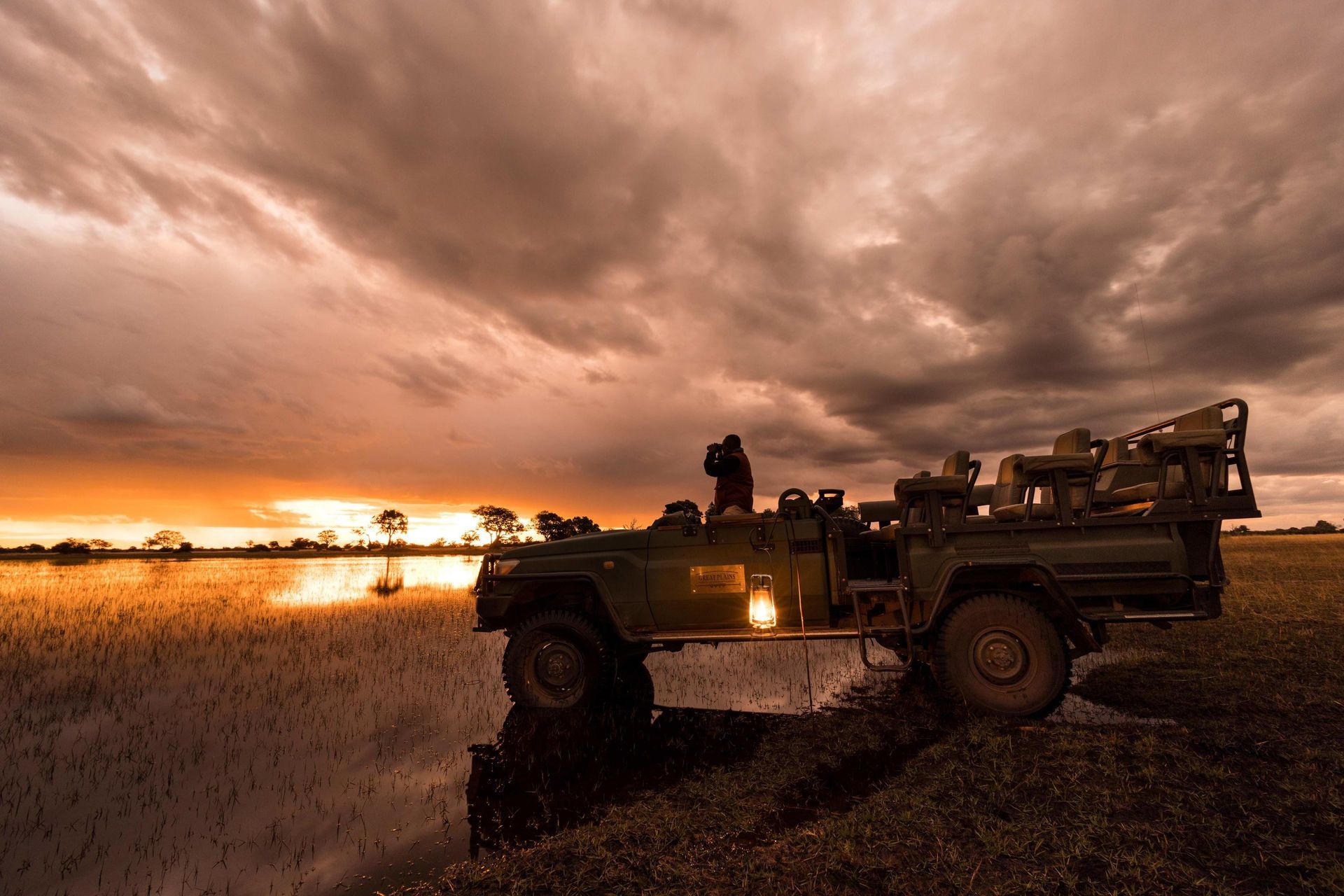 Safari vehicle on waterlogged terrain at sunset; person taking photos.
