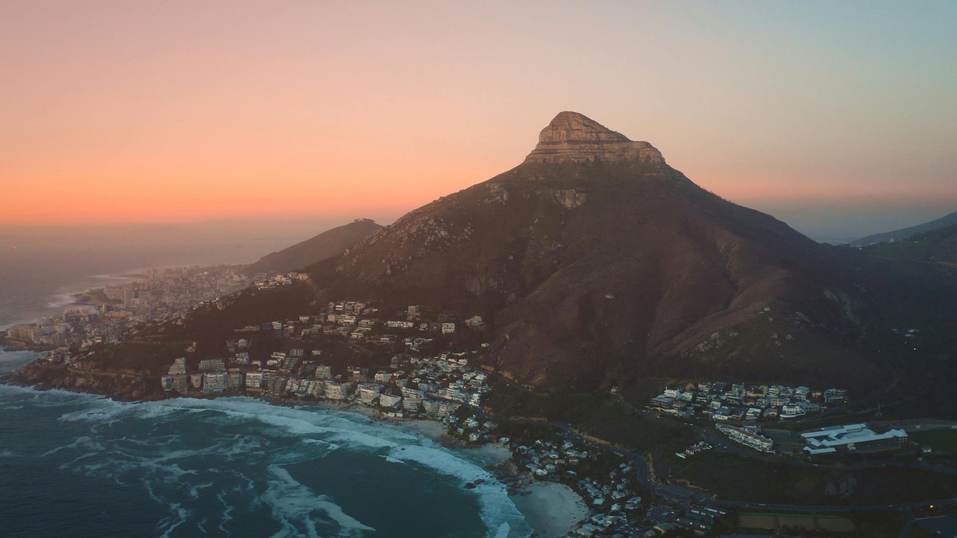 Lion's Head mountain at sunset, overlooking coastal town and ocean. Warm orange and blue hues.