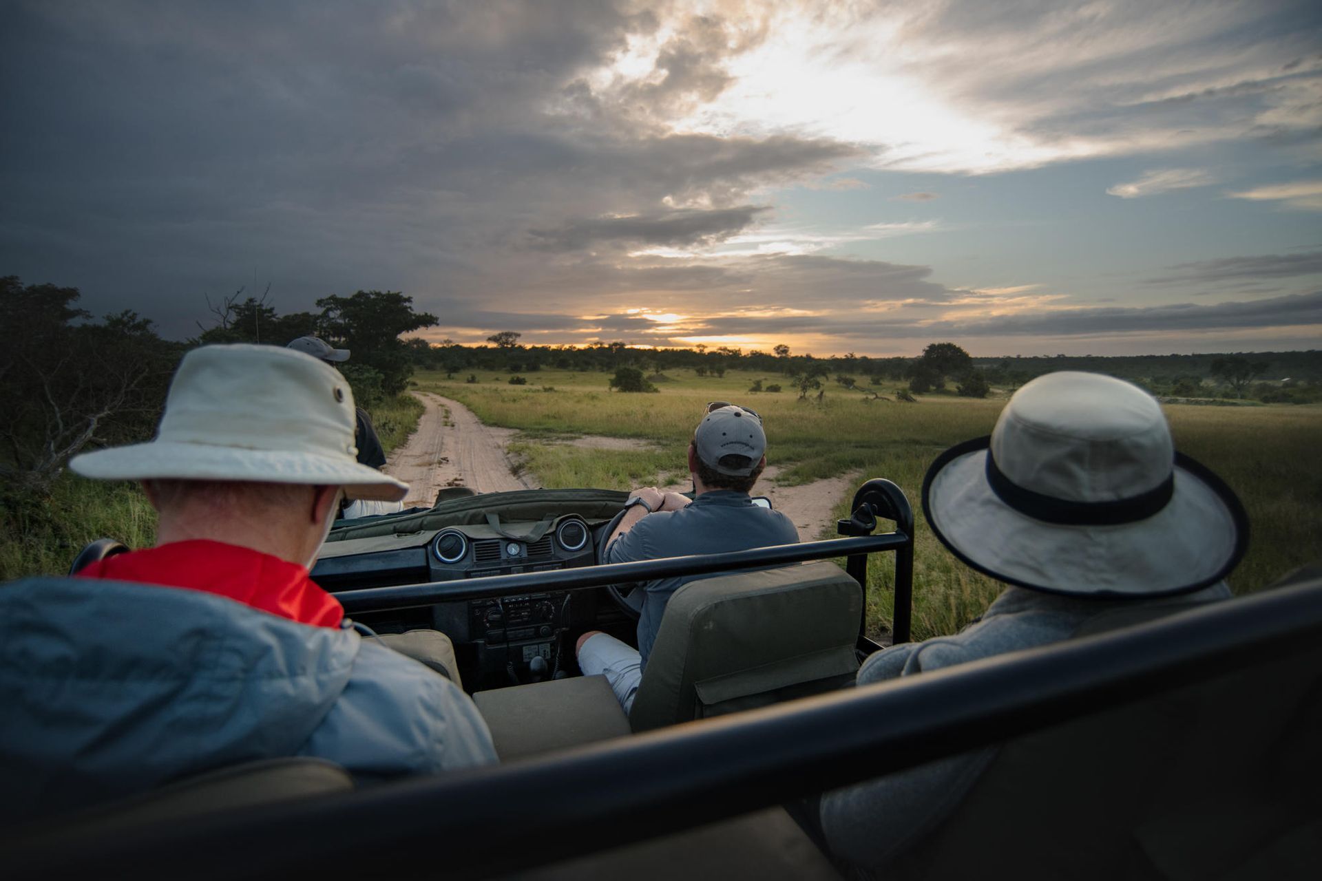Safari vehicle, three people, driving through savanna at sunset.