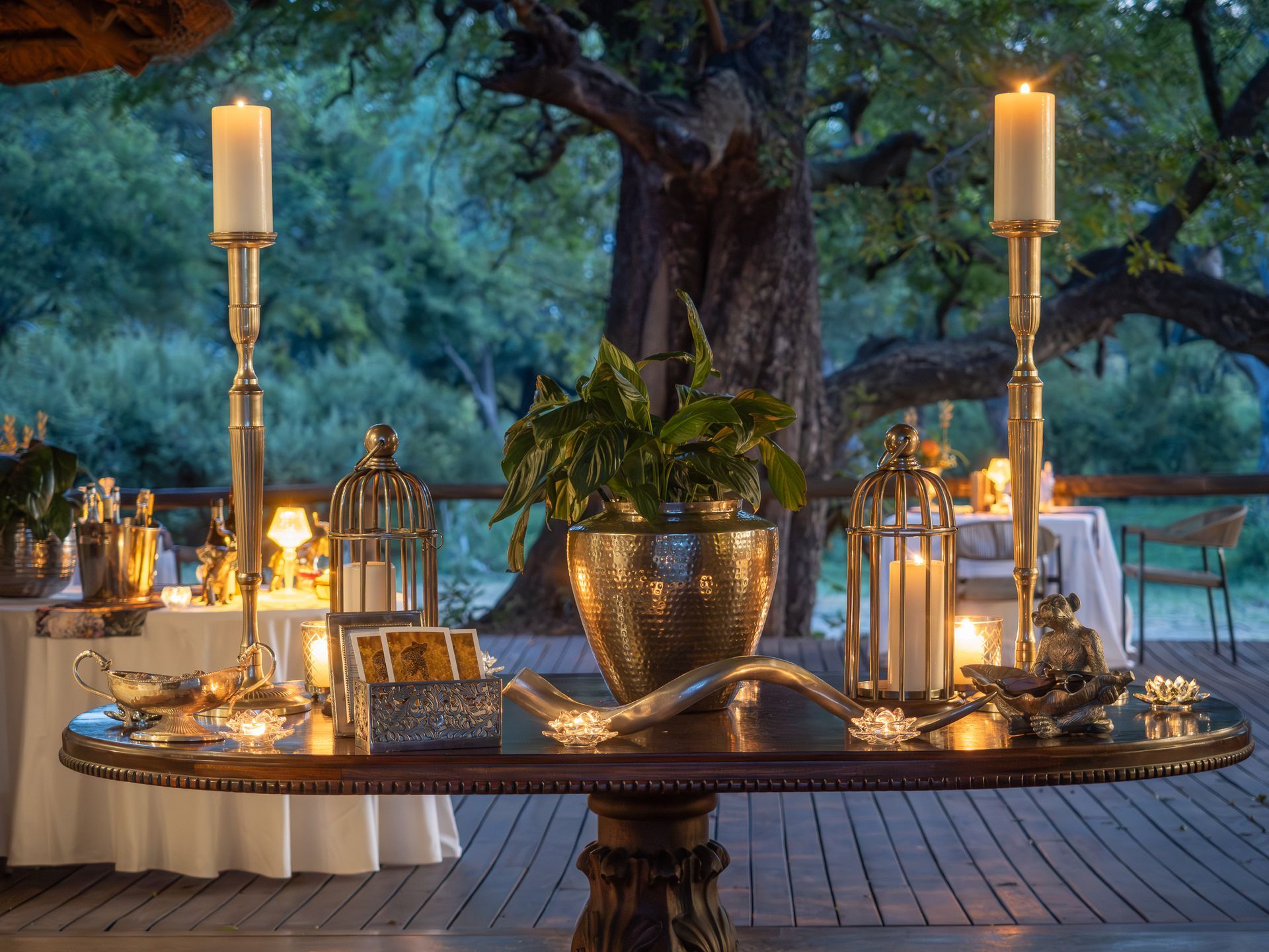 A candlelit outdoor table with decorations, including two tall candlesticks and a plant, in a forest setting.