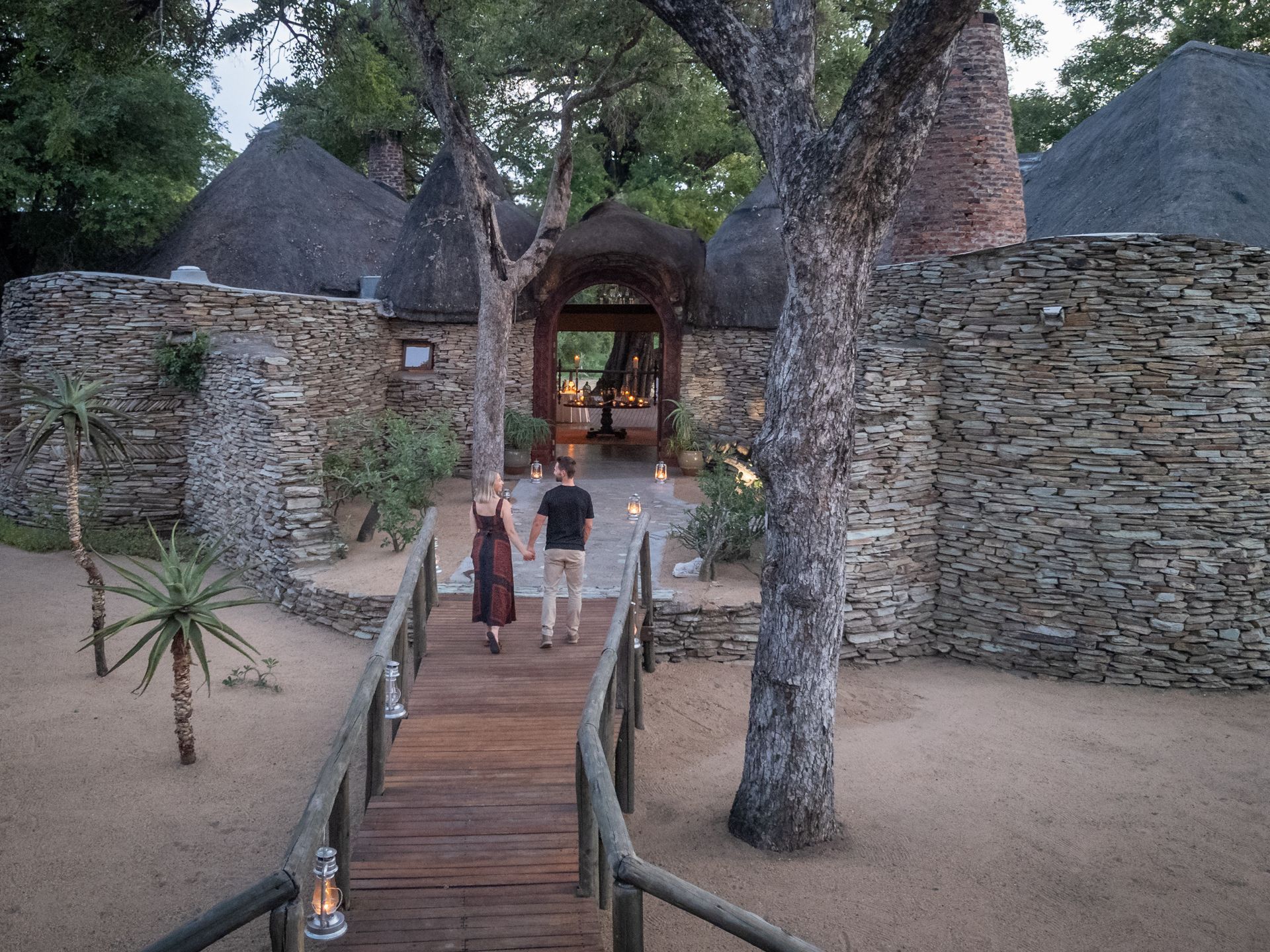 Couple walks along a wooden bridge towards an outdoor dining area at a stone and thatch building.