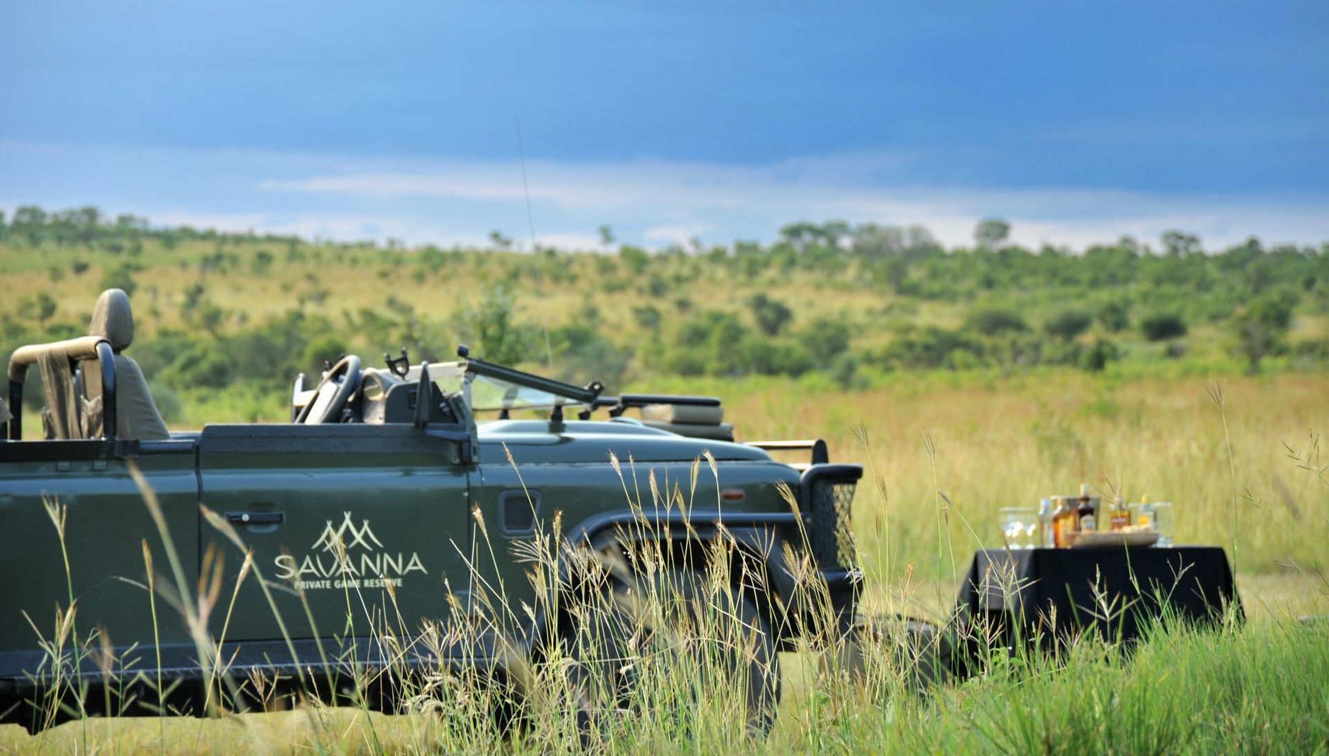 Safari vehicle parked next to a table set for a meal, with tall grass and a savanna landscape.