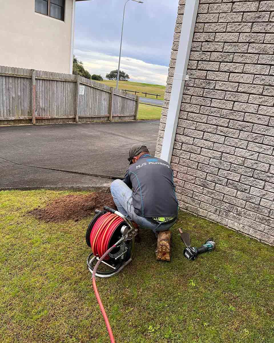 A Man Is Kneeling In The Grass Next To A Brick Wall — Todd Williams Plumbing and Gas Fitting In Forster, NSW