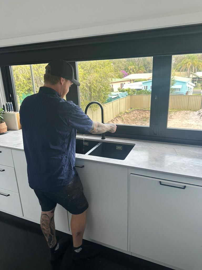 A Man Is Standing Next To A Sink In A Kitchen — Todd Williams Plumbing and Gas Fitting In Forster, NSW