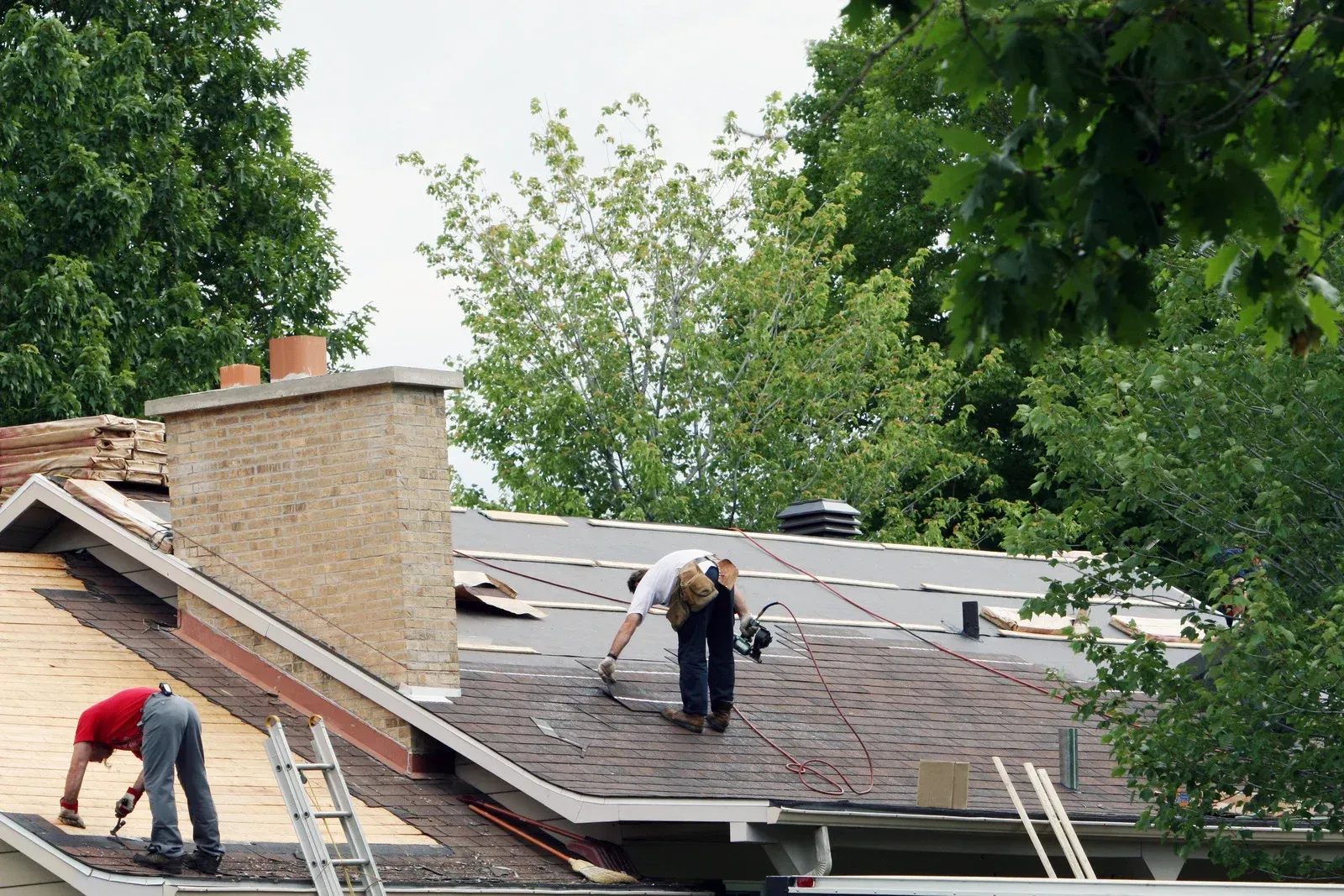 Workers repairing a shingled roof beside a brick chimney, surrounded by trees