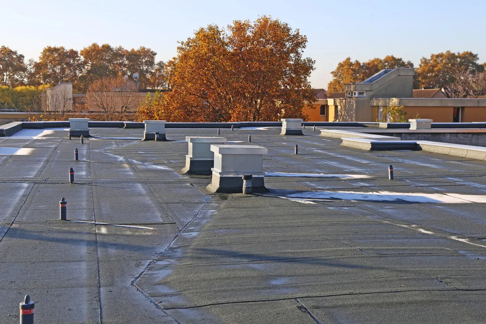 Flat rooftop with vents and patches of snow under autumn trees and a clear sky