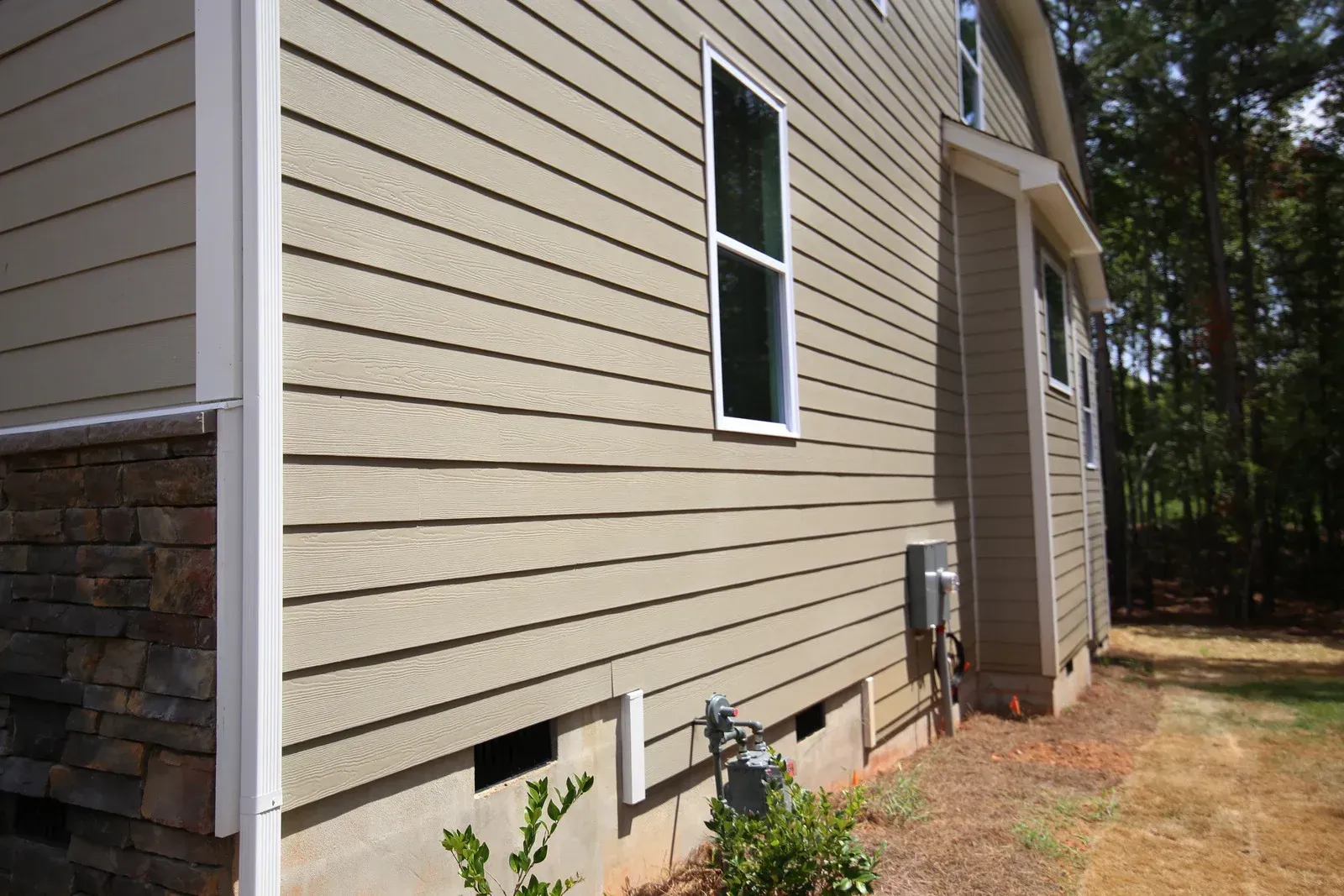 Side view of a beige house with stone siding detail and a window, beside a wooded yard.