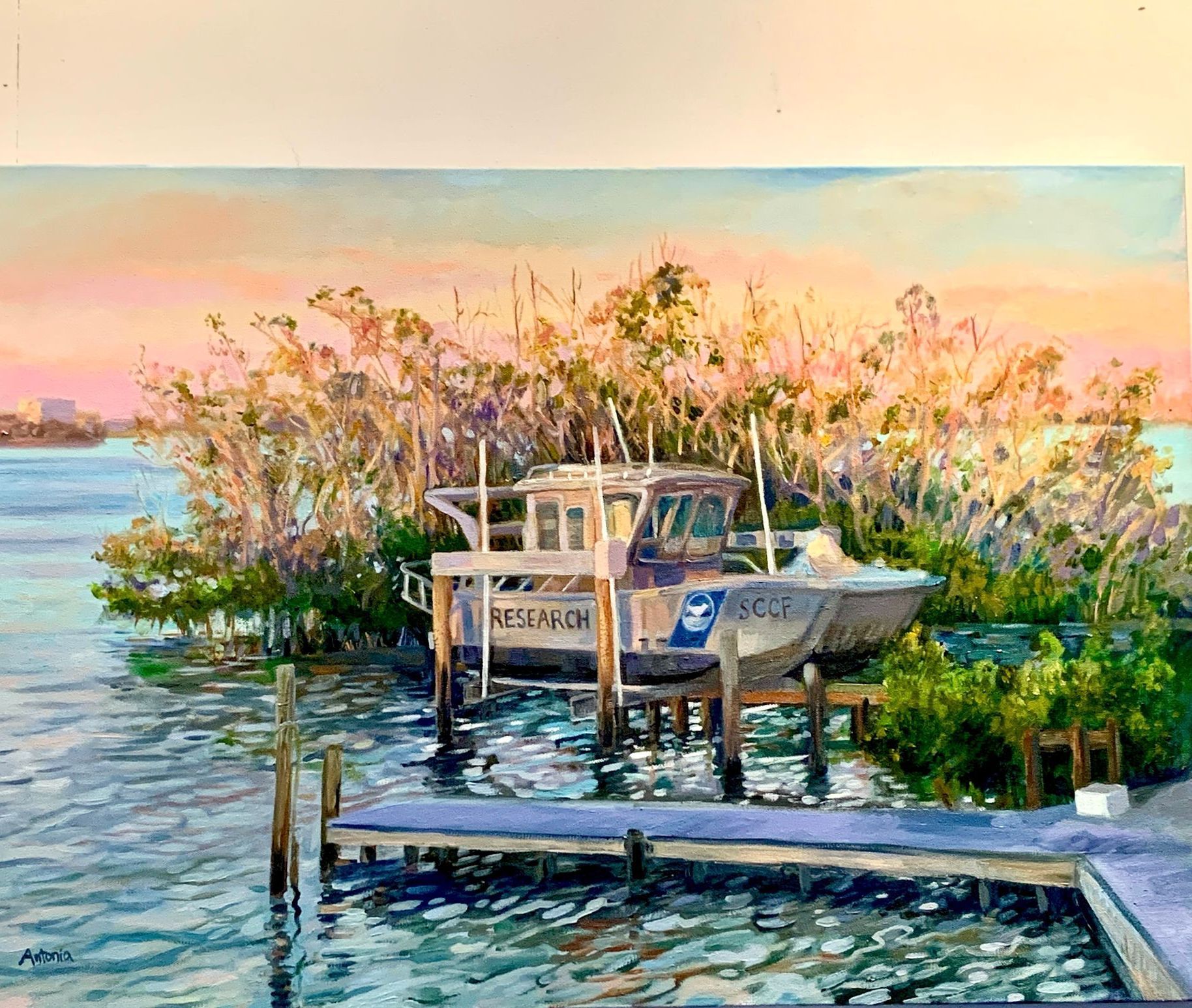 Boat on a lift beside a dock, in front of trees, at sunset. Calm water.