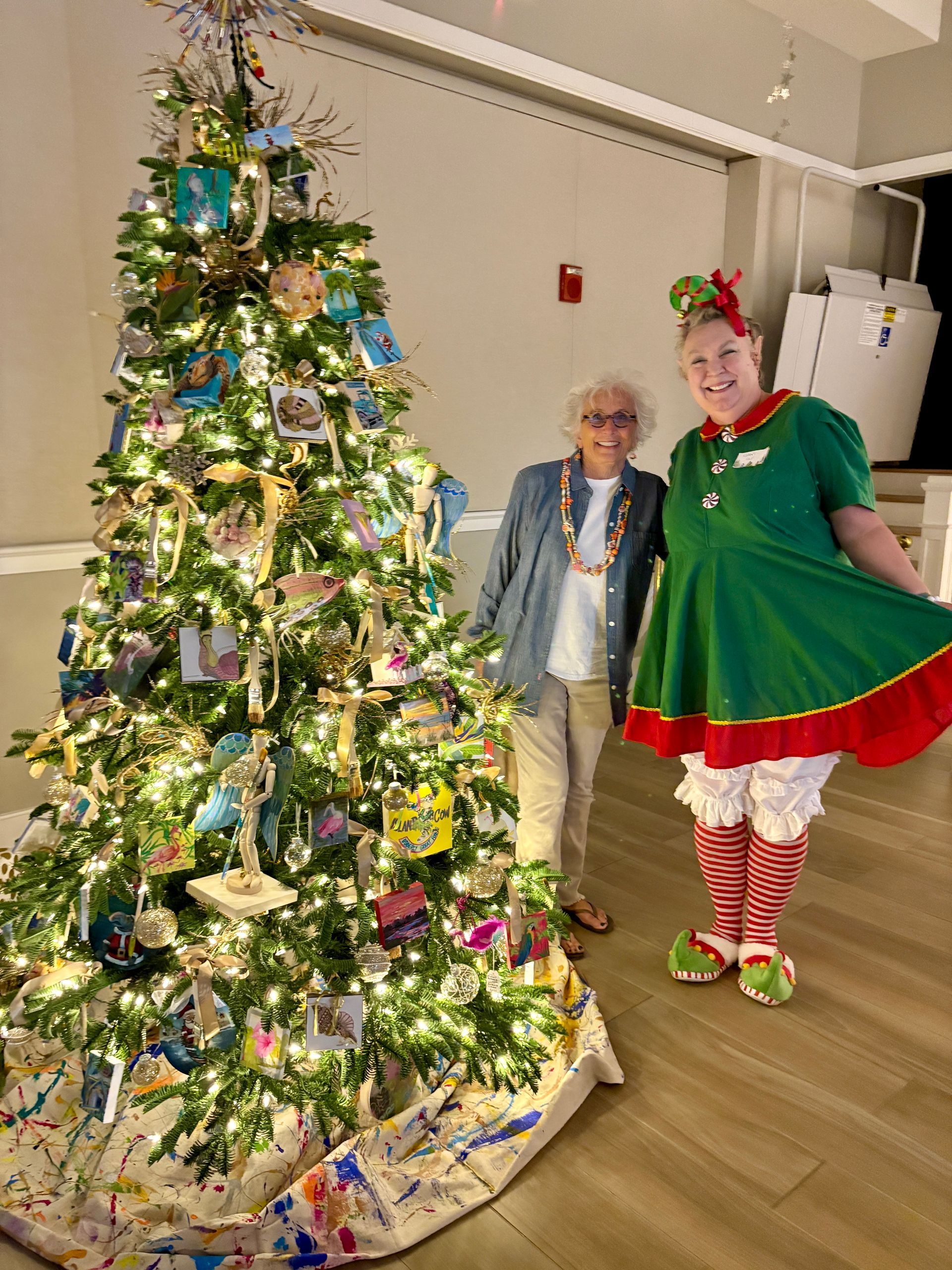 Woman in elf costume poses with another person by a decorated Christmas tree.
