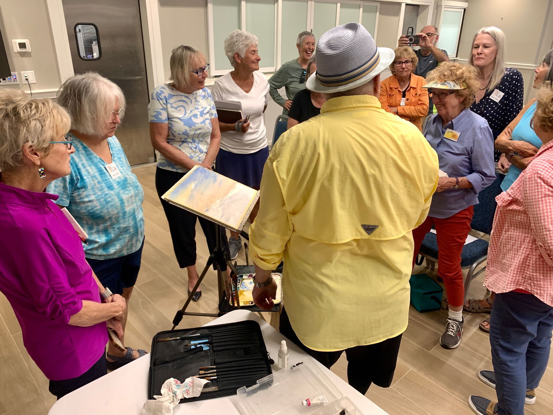 A man in a yellow shirt demonstrates painting to a group of women in a workshop.