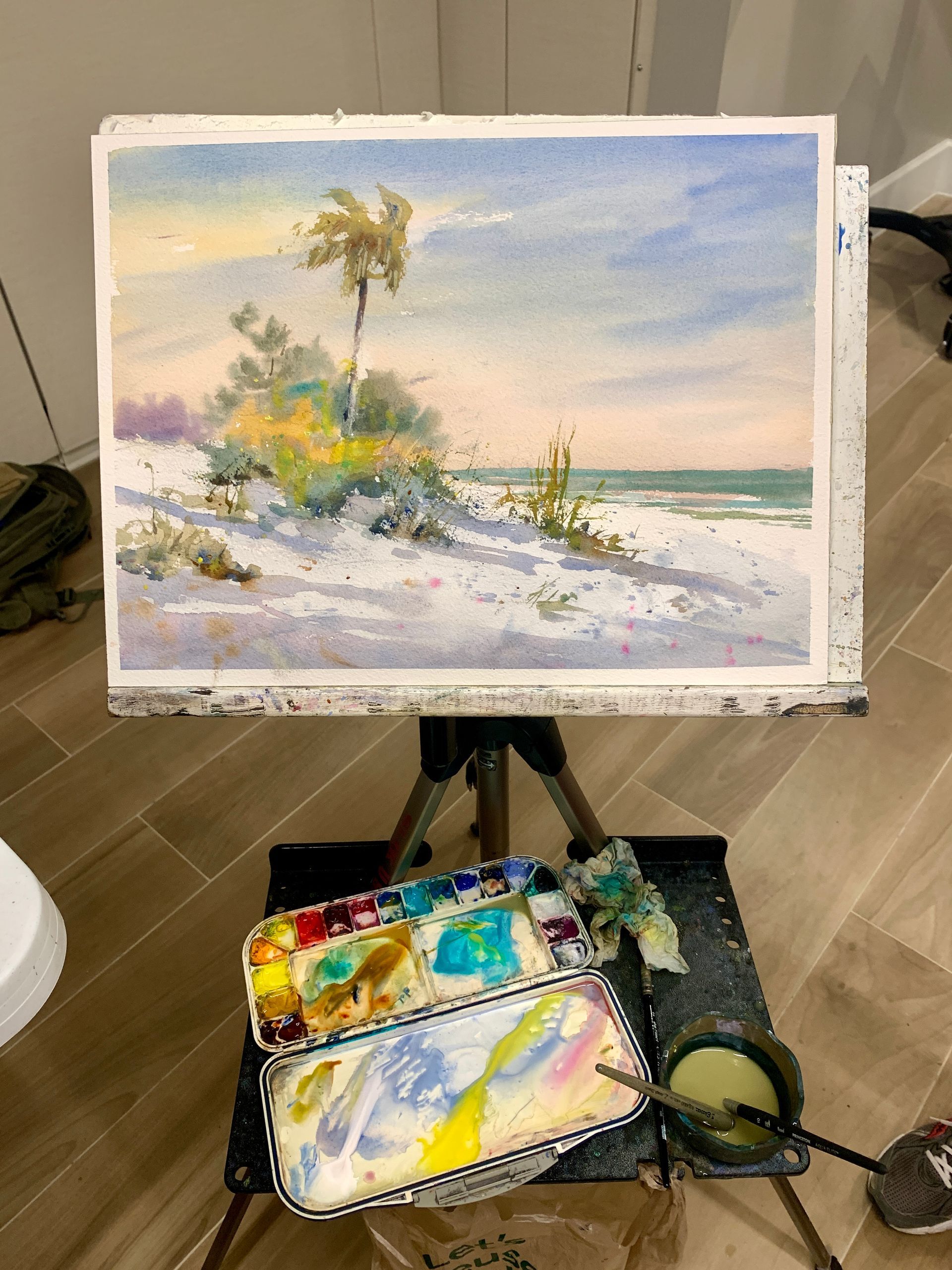 Watercolor painting on an easel of a beach scene: sand dunes, palm tree, and palette of paints.