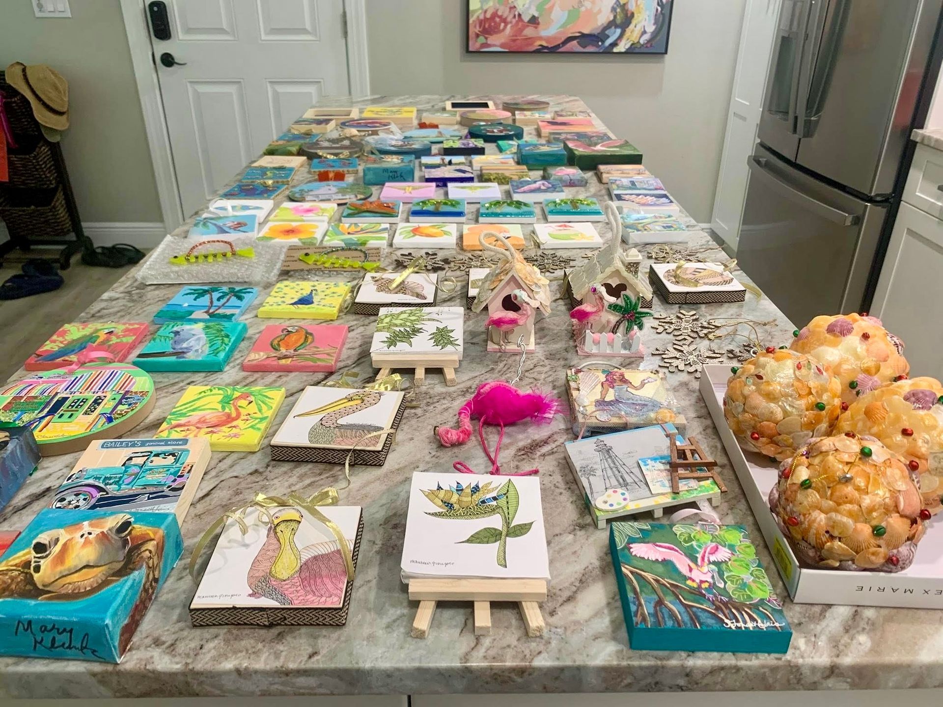 A kitchen countertop covered with numerous colorful paintings and small easels, ready for auction.