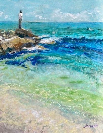 Person stands on rocks, facing vibrant turquoise ocean. Clear, rippling water in foreground, blue sky.