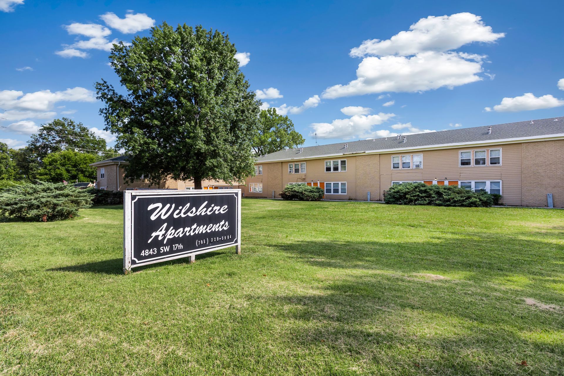 Wilshire Apartments sign in front of a two-story brick building, on a grassy lawn under a blue sky.