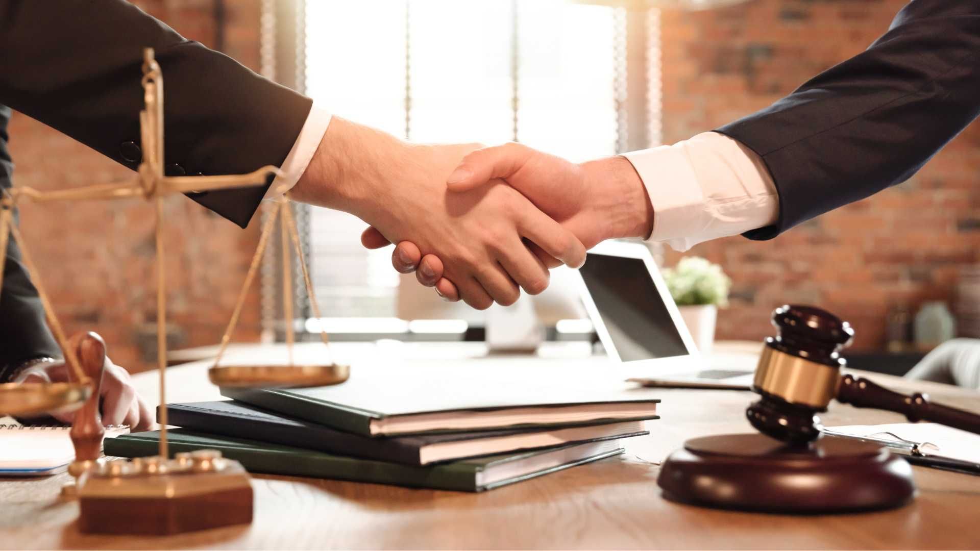 Two people shaking hands over a desk with law books, a gavel, and scales in a legal office.