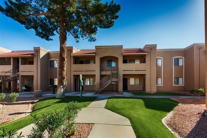 Apartment complex with tan buildings, green lawn, and a sidewalk under a clear blue sky