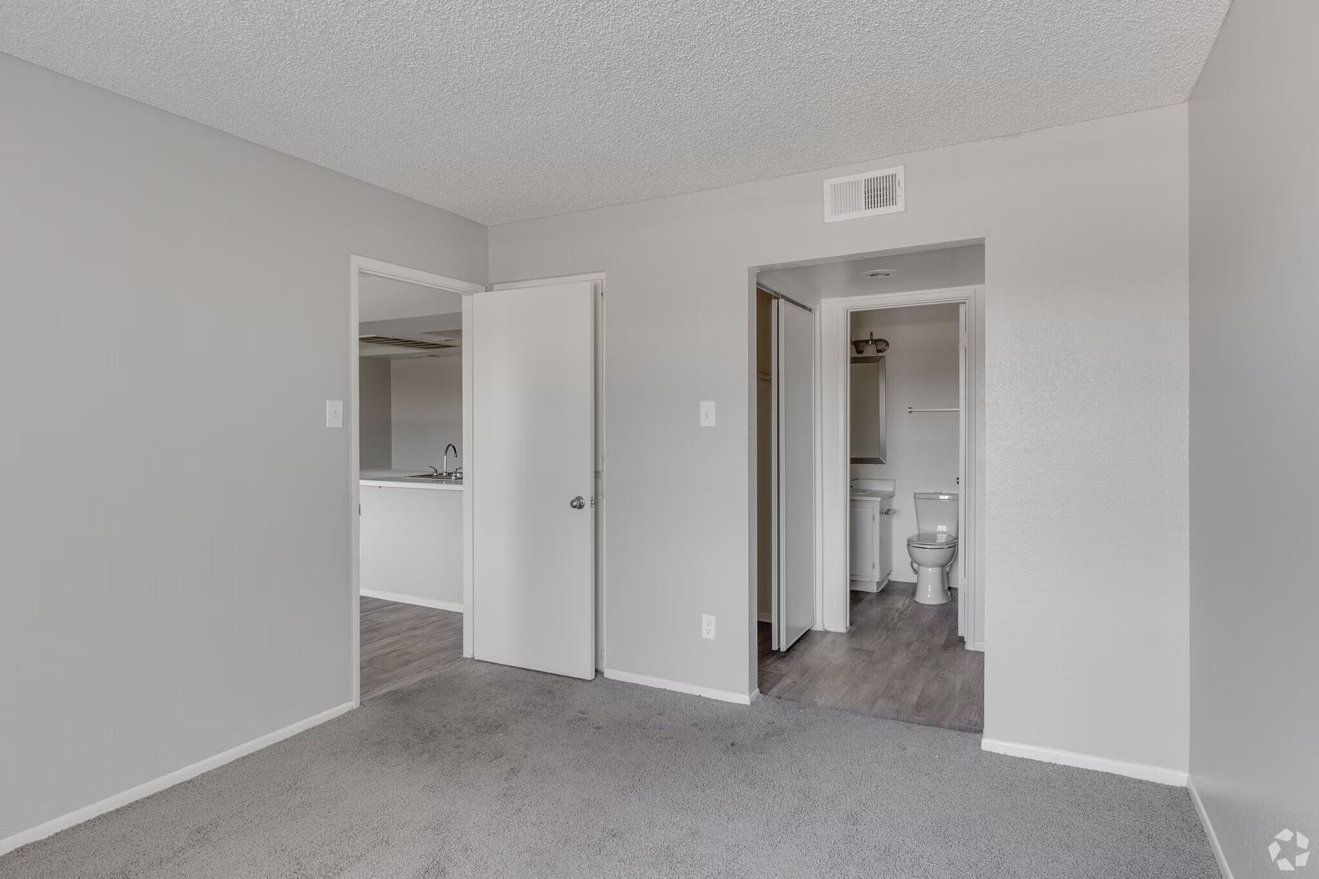Empty beige bedroom with carpet, open doors, and a bathroom visible through one doorway