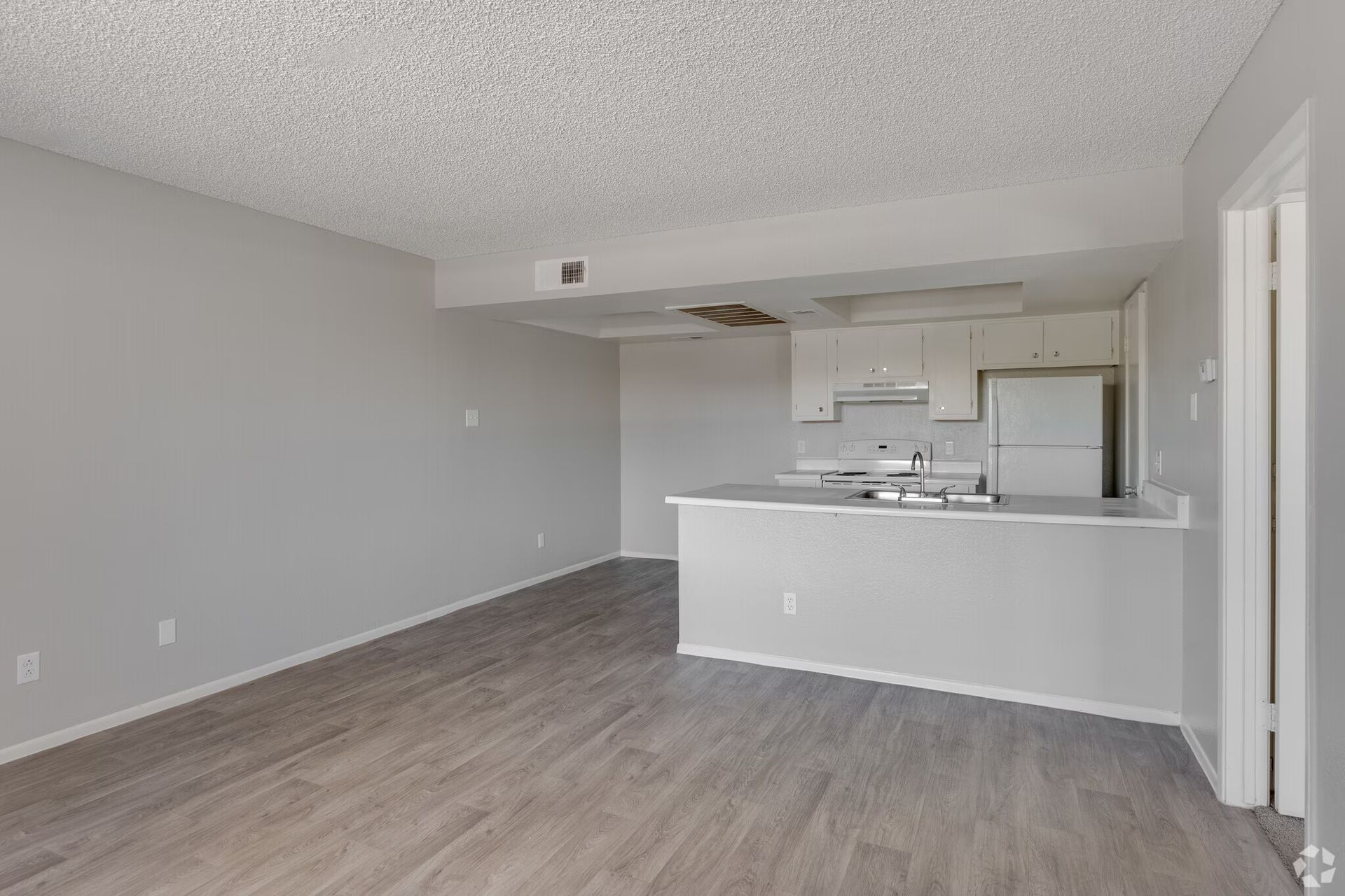 Empty living room with gray walls and wood floors, opening to a kitchen with a white counter.