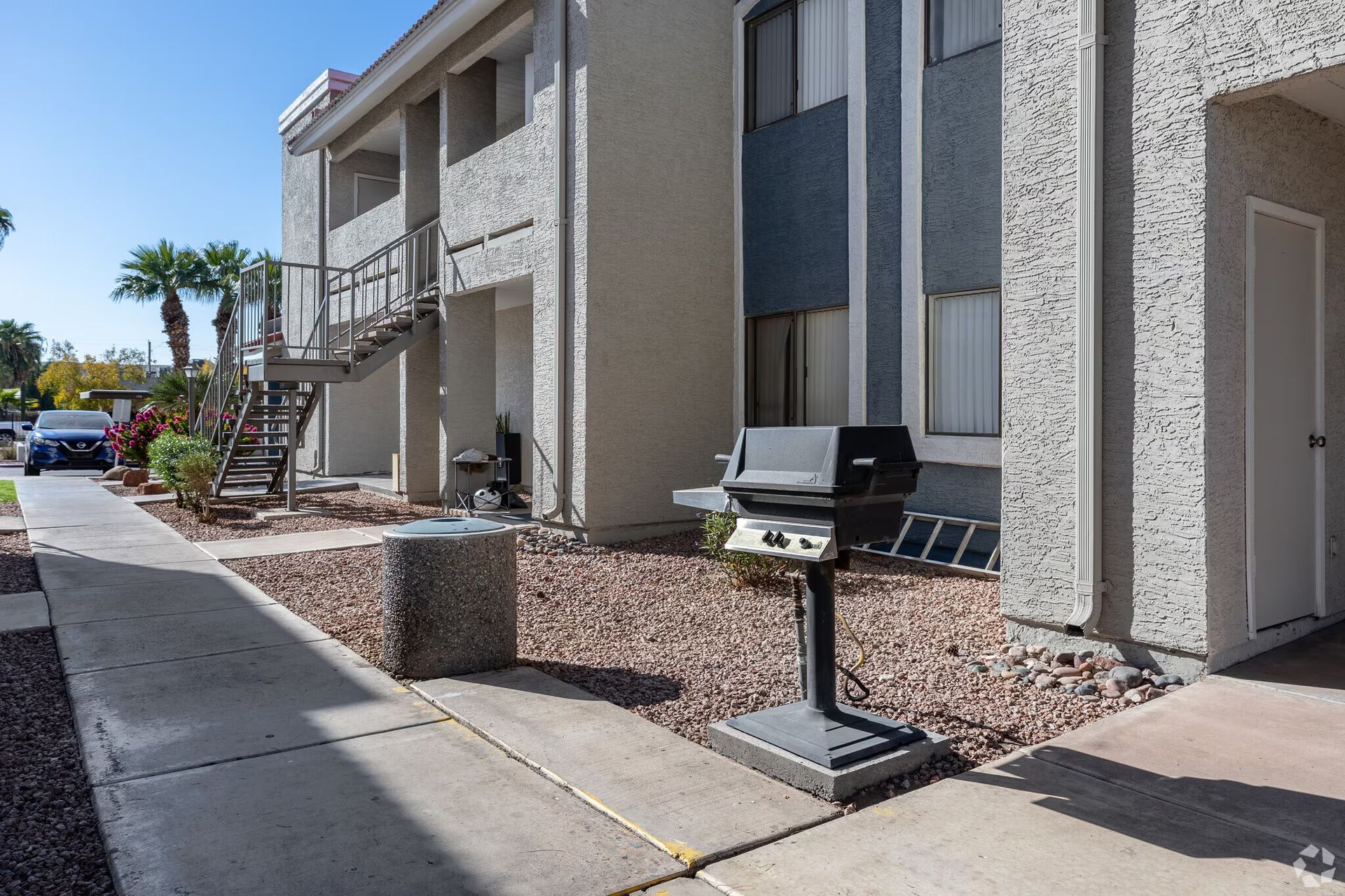 Apartment walkway with a barbecue grill and desert landscaping beside stucco buildings.