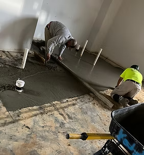 Two construction workers leveling wet concrete on a floor. One kneels, the other uses a screed.