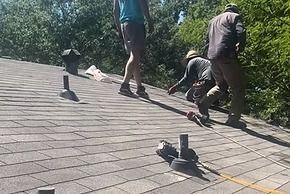 Three people on a rooftop, likely repairing or inspecting it. The roof is covered in shingles, and there are vents.