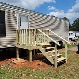 Mobile home with wooden steps and railing. The steps lead to a white door. A truck is parked nearby on a grassy lot.