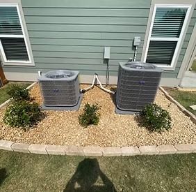 Two air conditioning units on a bed of tan gravel with small green plants, set against a light green house wall.