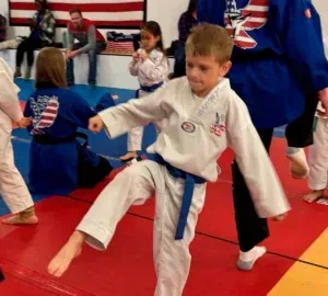 Child practicing a martial arts kick in a dojo, wearing a white uniform with a blue belt.