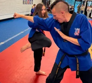 Martial arts instructor in blue gi demonstrating a kick with a student on red and blue mats.